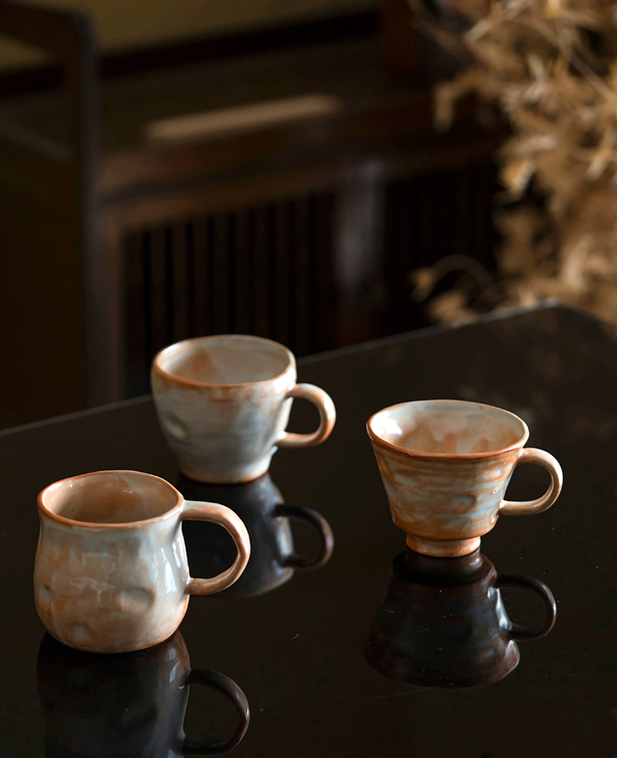 three handcrafted ceramic coffee and tea cups in the clayshade collection with warm terracotta and ash glaze, arranged on a reflective surface in a serene setting