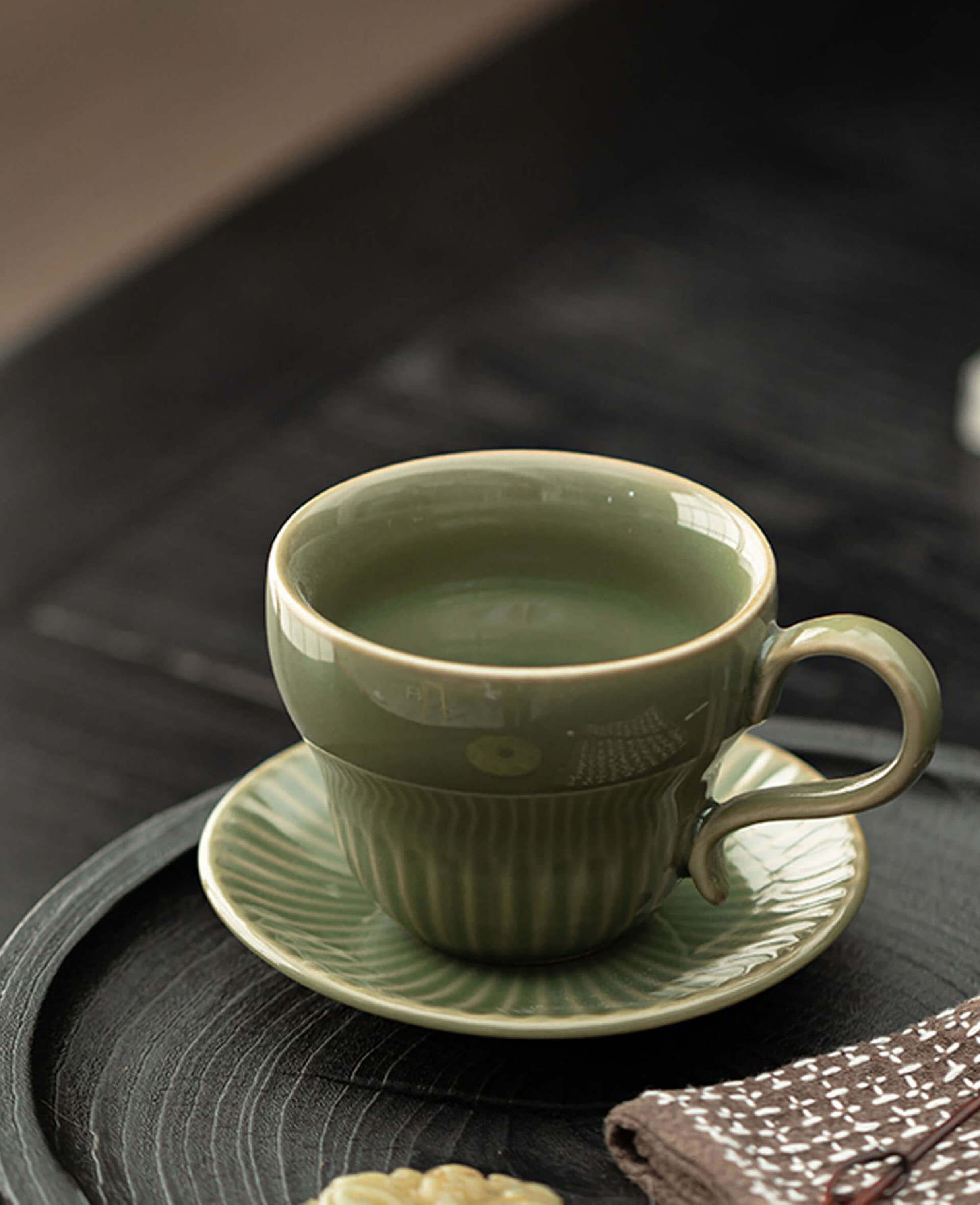 greenstill crest porcelain tea and coffee cup with fluted design resting on a matching saucer atop a black wooden tray beside sweet treats