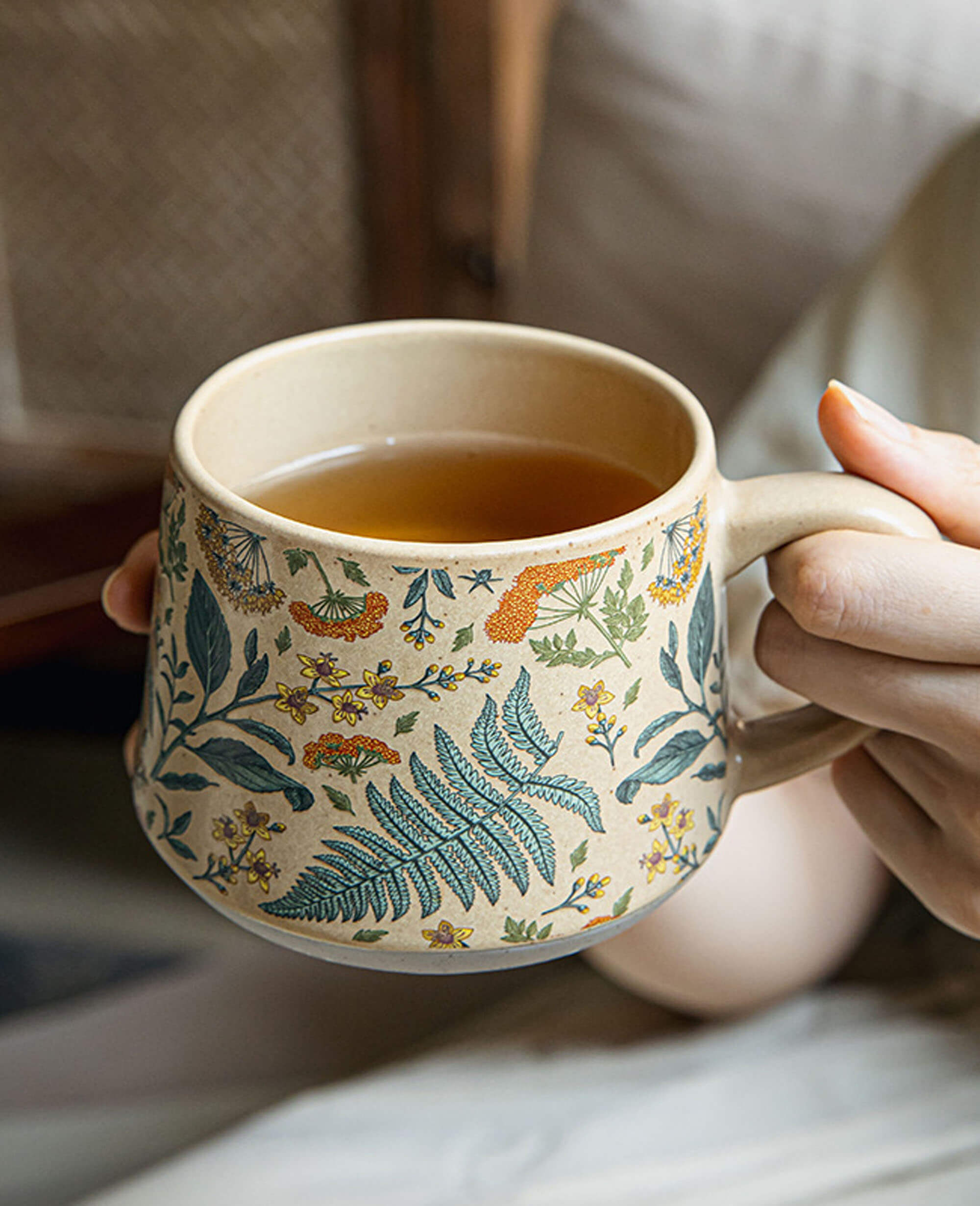 hand holding the july note floral ceramic mug with tea showcasing the curved handle and botanical design