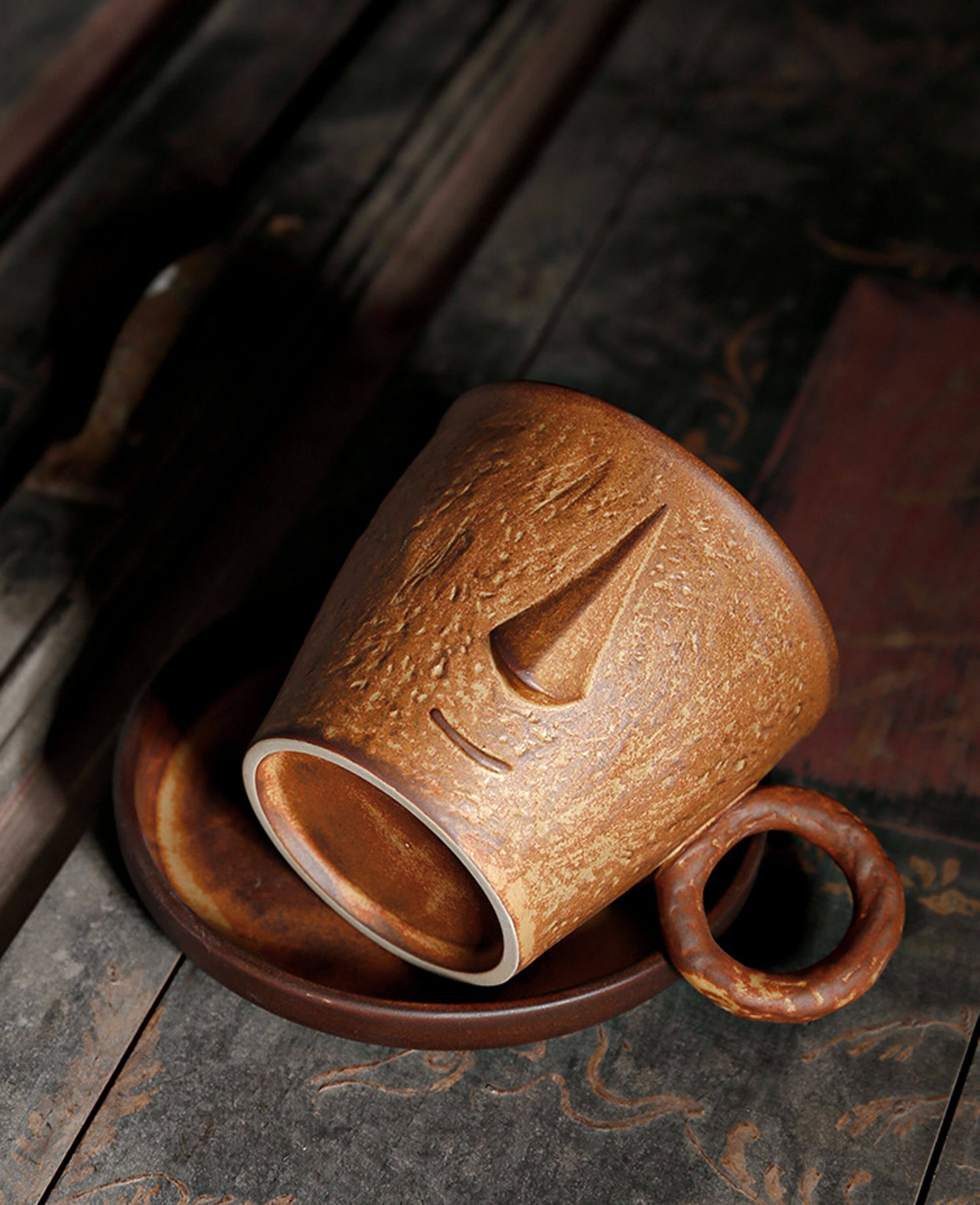 toffee reverie muse mug laying on its side revealing the sculpted facial features and rustic ring handle paired with a saucer
