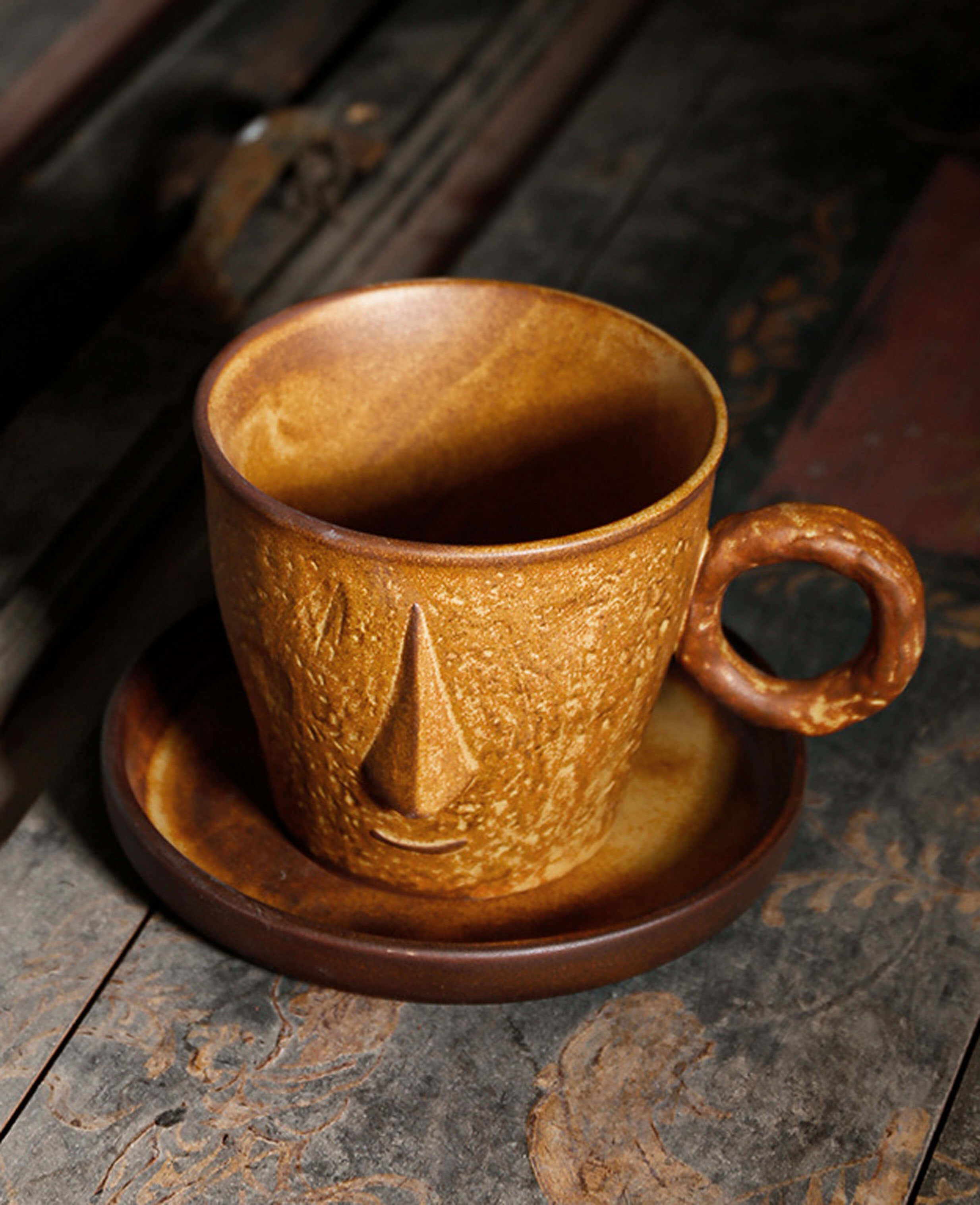 toffee reverie muse coffee mug with sculpted face design sitting on a matching saucer on a vintage wooden surface