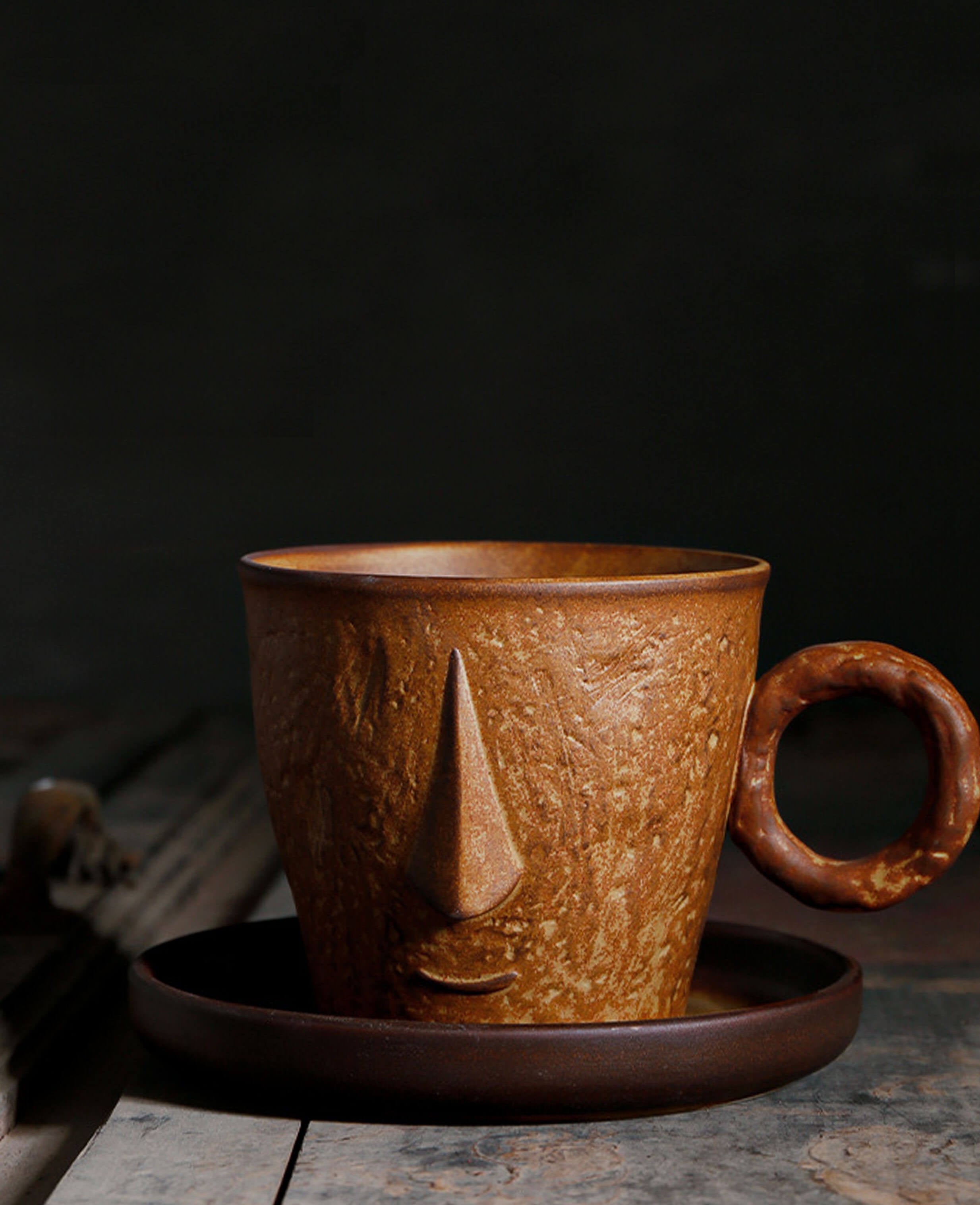 front facing toffee reverie muse mug with artistic face motif on dark background and vintage wood setting