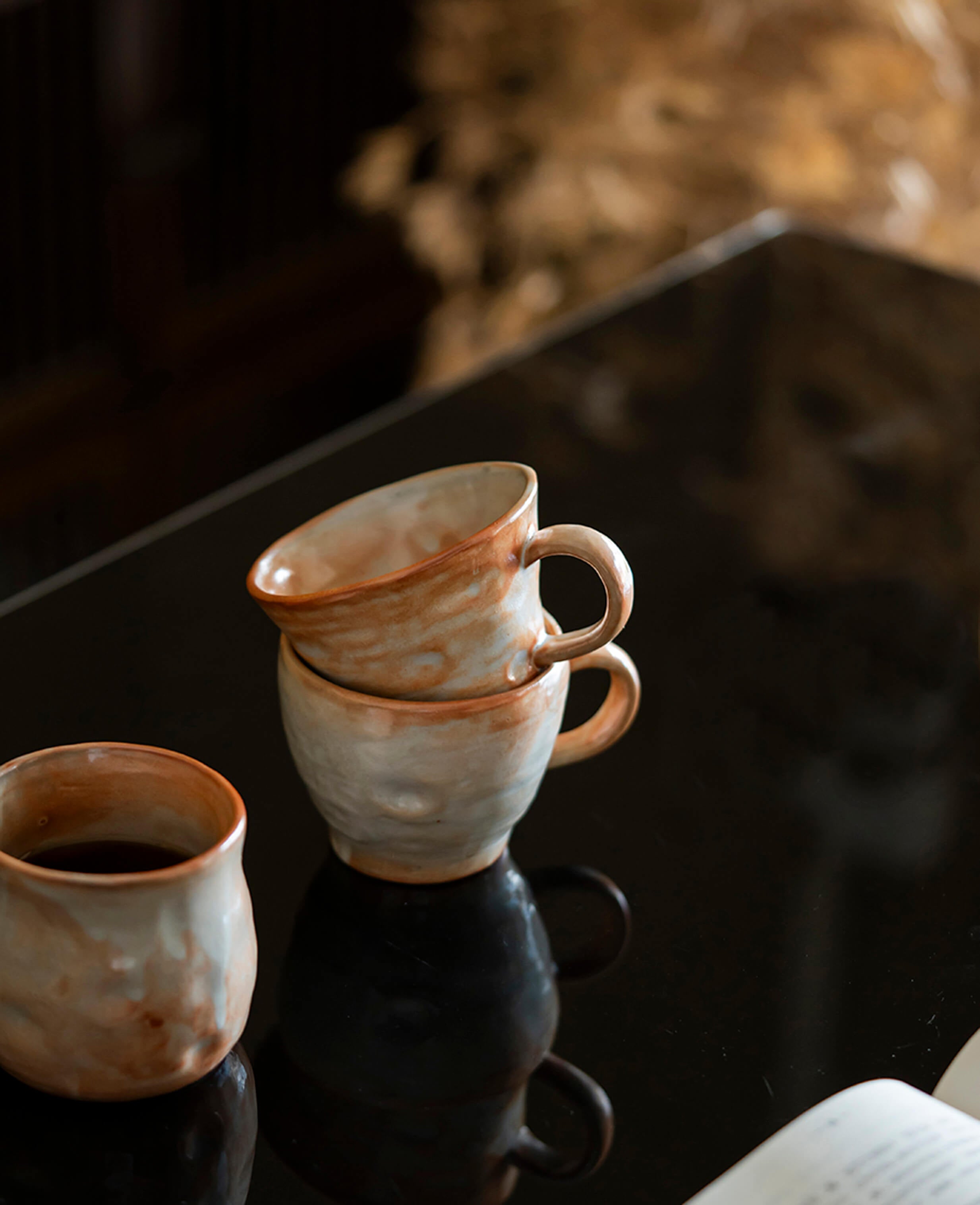 stacked handcrafted ceramic coffee and tea mugs from the clayshade collection with ash grey and terracotta glaze resting on a glossy surface beside an open book in a quiet atmosphere