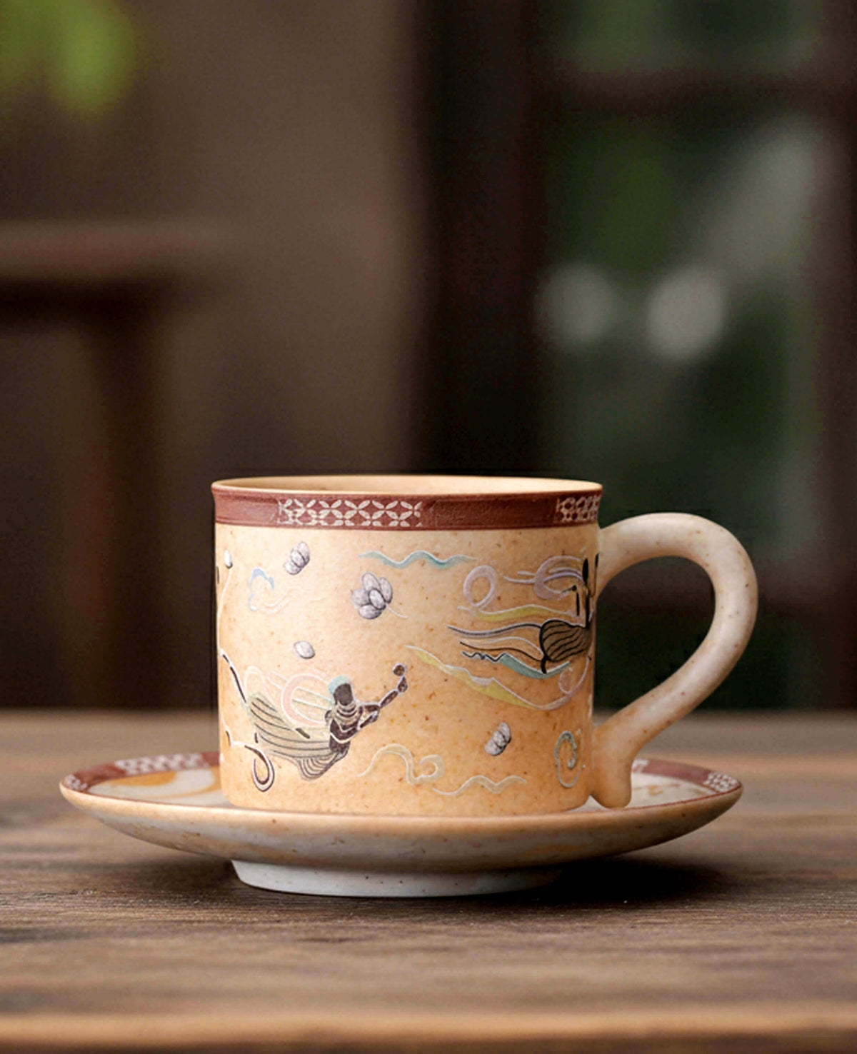 A beige ceramic coffee cup and saucer set with hand-painted traditional motifs, including a figure and flowing lines, set on a rustic wooden table with a blurred background.