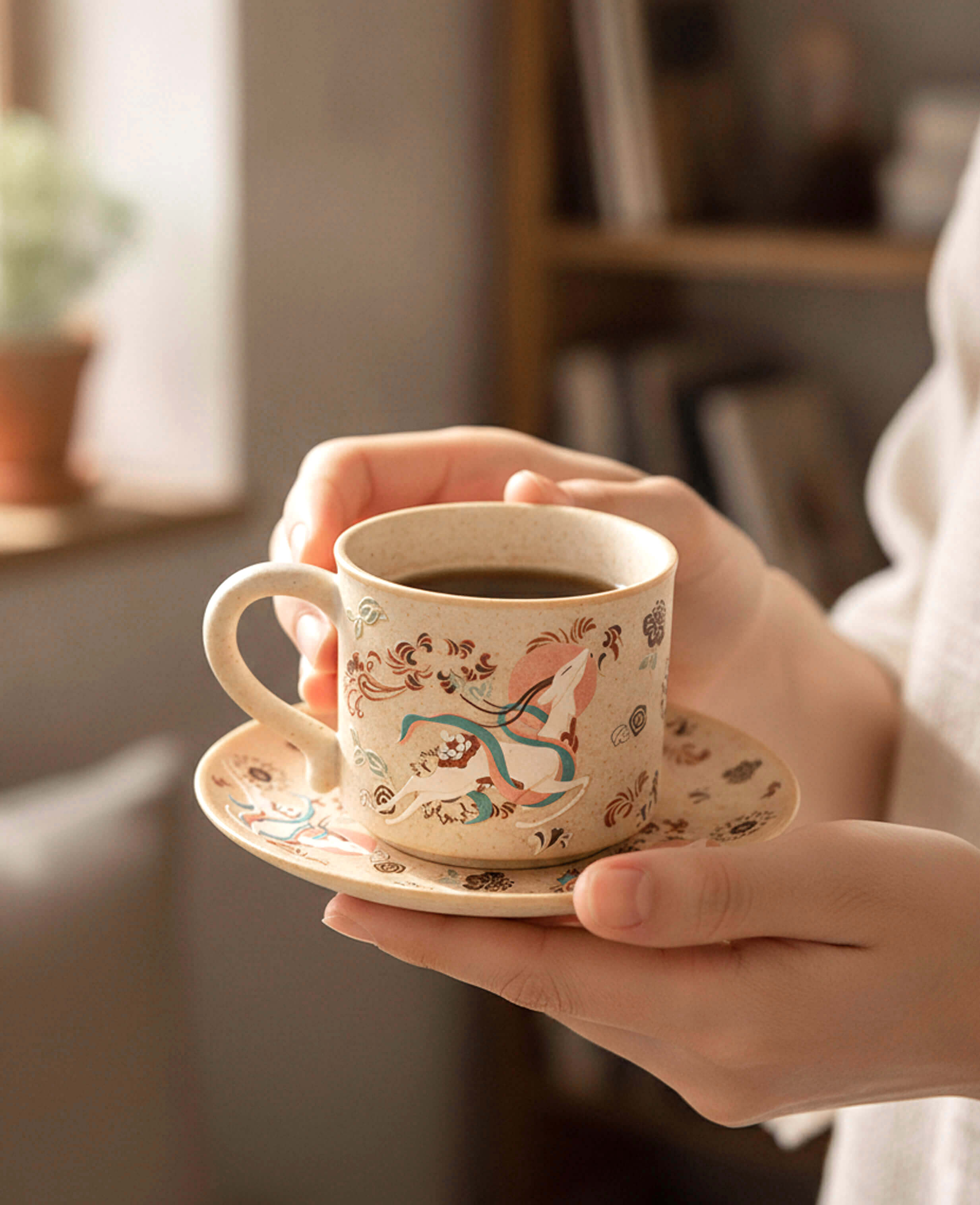 A person holds a ceramic cup and saucer decorated with a nine coloured deer illustrated design, containing dark coffee, in a softly lit indoor setting.
