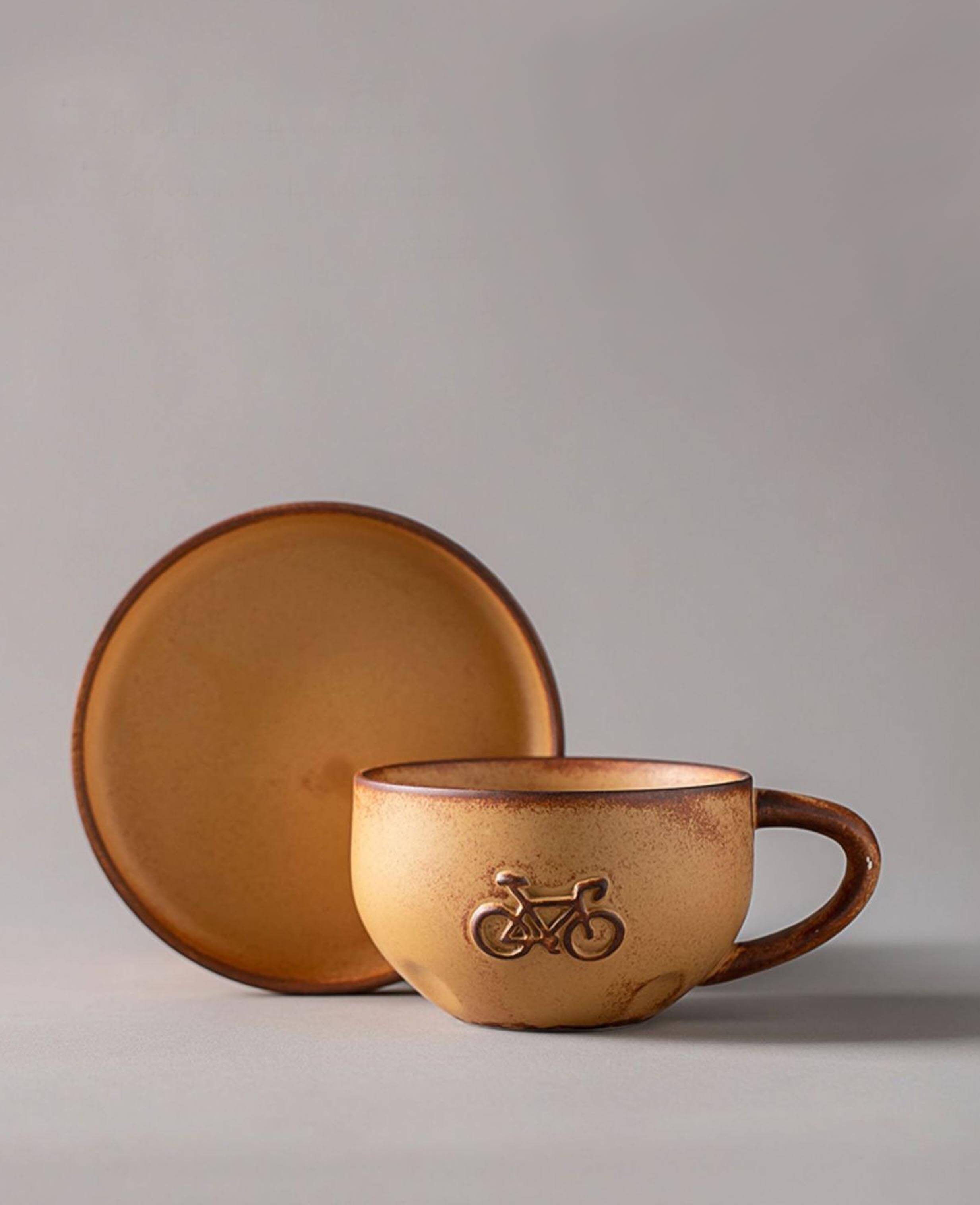 handcrafted stoneware rustic coffee mug pedalstone with bicycle relief and saucer on grey background