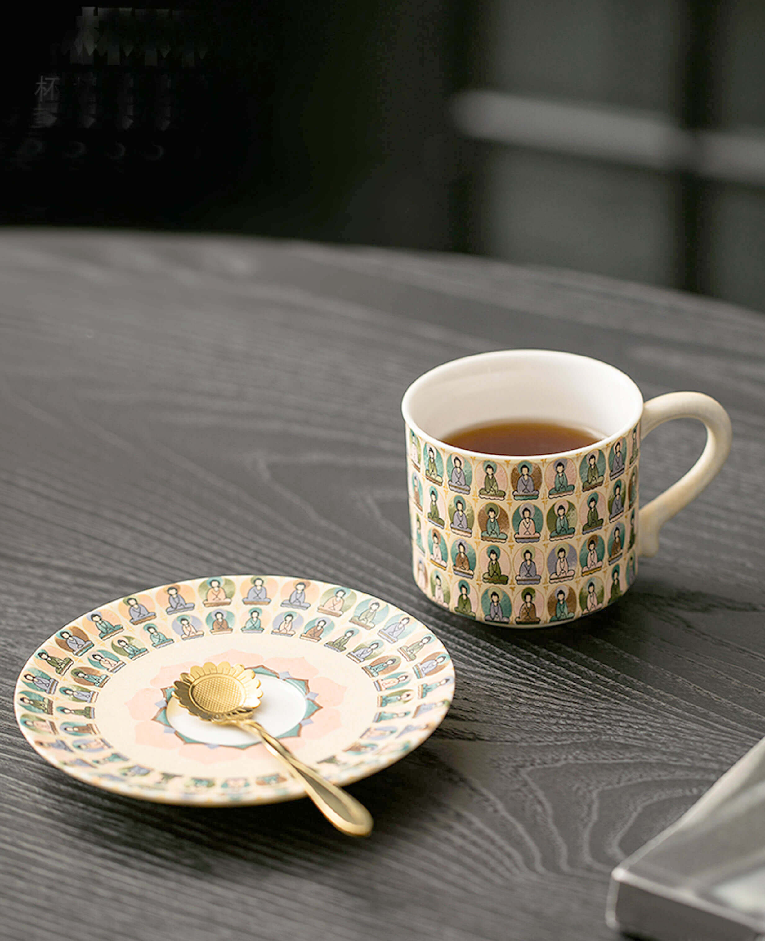 ceramic cup and saucer set with pastel thousand buddhas pattern styled with golden spoon on textured black table