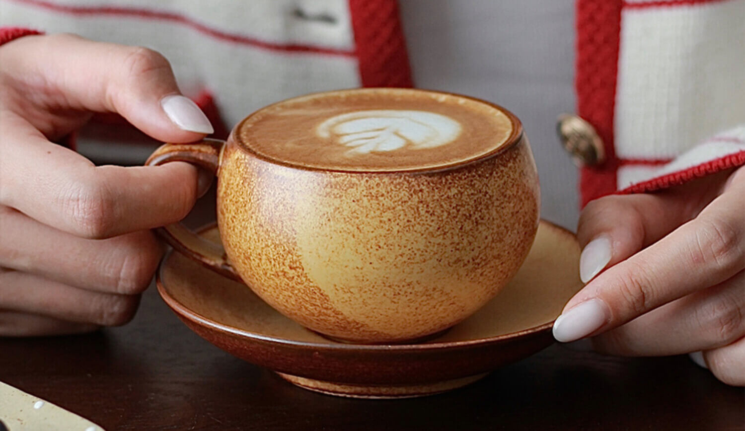 hands holding a warm cup of latte in the toffee glaze ovolo ceramic cup and saucer featuring rich amber tones and frothy art