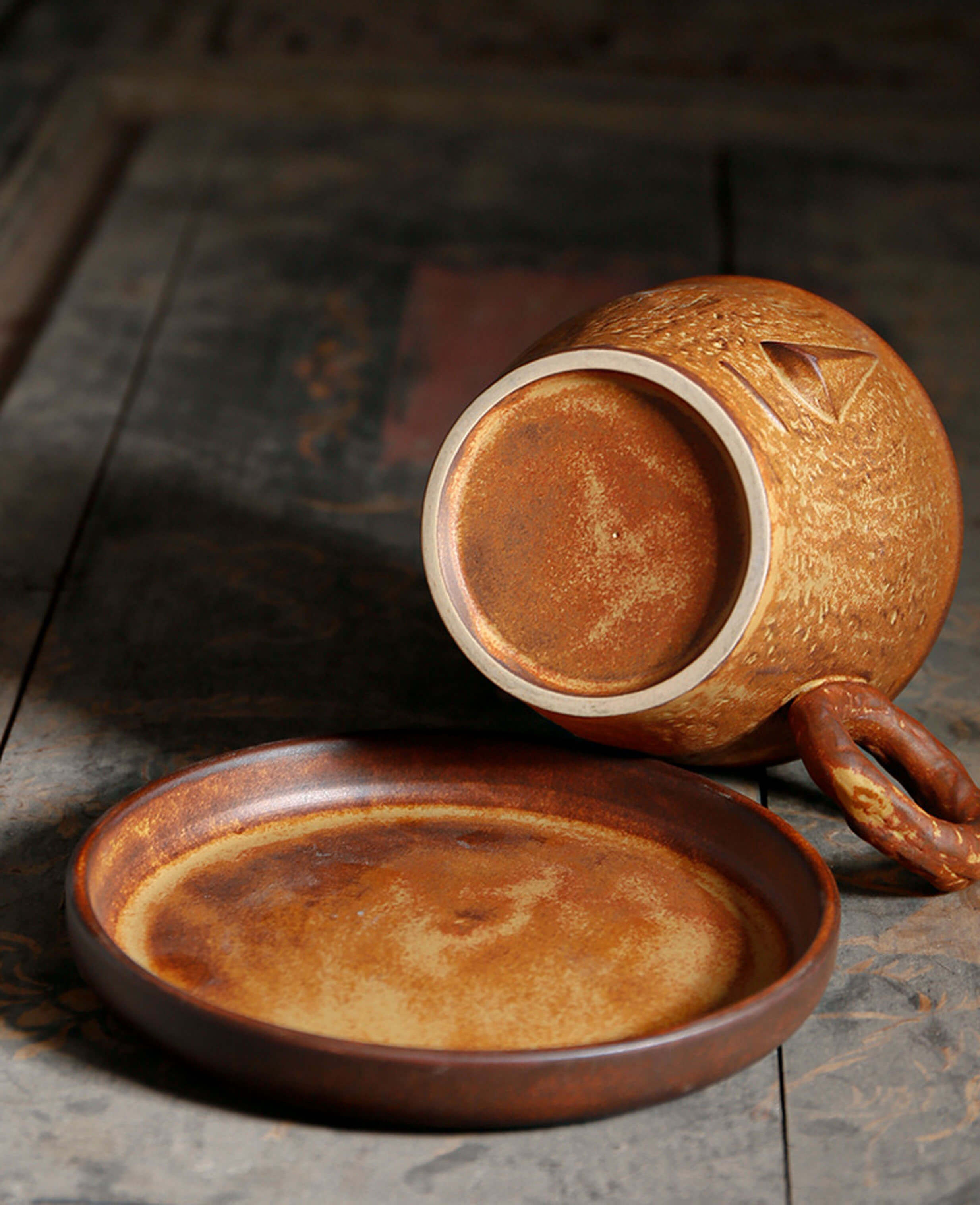 bottom view of toffee reverie muse coffee mug with sculpted face, placed beside matching saucer on distressed wooden tabletop